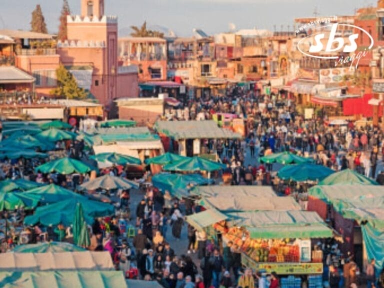 Vivace mercato all'aperto con numerose bancarelle coperte da pensiline verdi, circondato da una grande folla di persone nella vivace Città Imperiali del Marocco. Edifici e negozi costeggiano lo sfondo sotto un cielo limpido, catturando l'essenza della tradizionale medine del Nord.