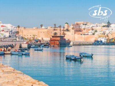 Vista di un porto con diverse piccole imbarcazioni in acqua, fortificazioni storiche sullo sfondo e un paesaggio urbano con edifici bianchi in cima a una collina. La scena esplode di colori che ricordano il Marocco. Logo "sbs viaggi" nell'angolo in alto a destra.