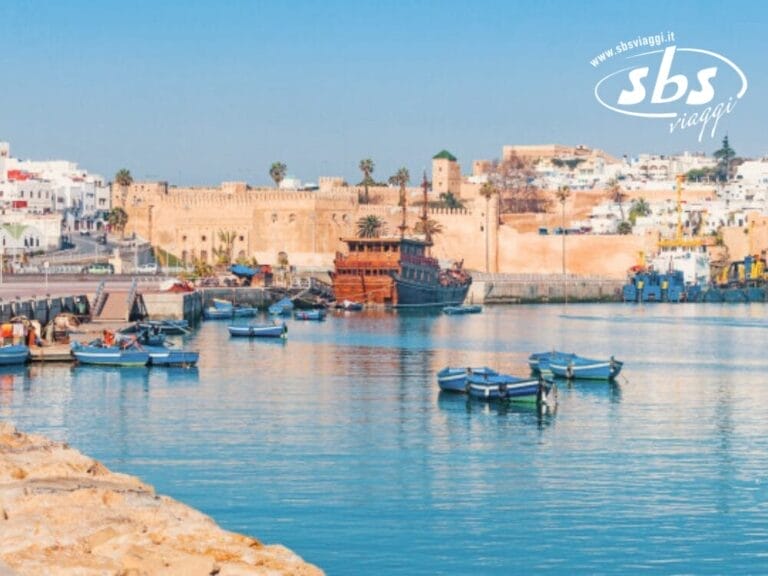 Vista di un porto con diverse piccole imbarcazioni in acqua, fortificazioni storiche sullo sfondo e un paesaggio urbano con edifici bianchi in cima a una collina. La scena esplode di colori che ricordano il Marocco. Logo 