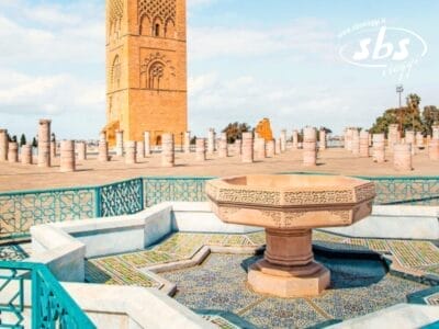 Una splendida fontana in pietra con intricate piastrelle adorna la facciata di un'alta torre, che ricorda la grandiosità di Marrakech. Colonne di marmo si ergono sparse sotto un cielo blu vibrante, riecheggiando la bellezza senza tempo della Città Imperiali del Marocco.