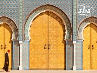 Durante il Gran Tour del Marocco, una persona in abiti tradizionali cammina con eleganza davanti a grandi e ornate porte dorate, impreziosite da intricati motivi a mosaico sull'affascinante facciata dell'edificio.