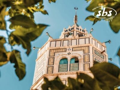 Una torre tunisina con piastrelle decorative e finestre ad arco, incorniciata da foglie verdi, sotto un cielo azzurro e limpido.