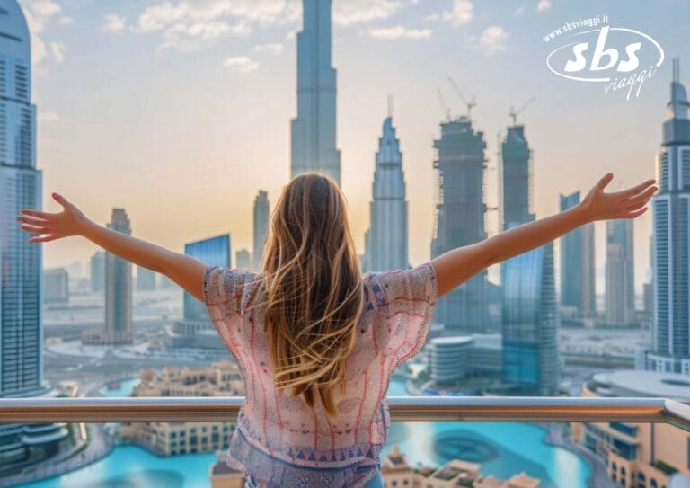 Una donna con i capelli lunghi è in piedi su un balcone a Dubai, con le braccia tese, mentre il sole tramonta dietro i moderni grattacieli.