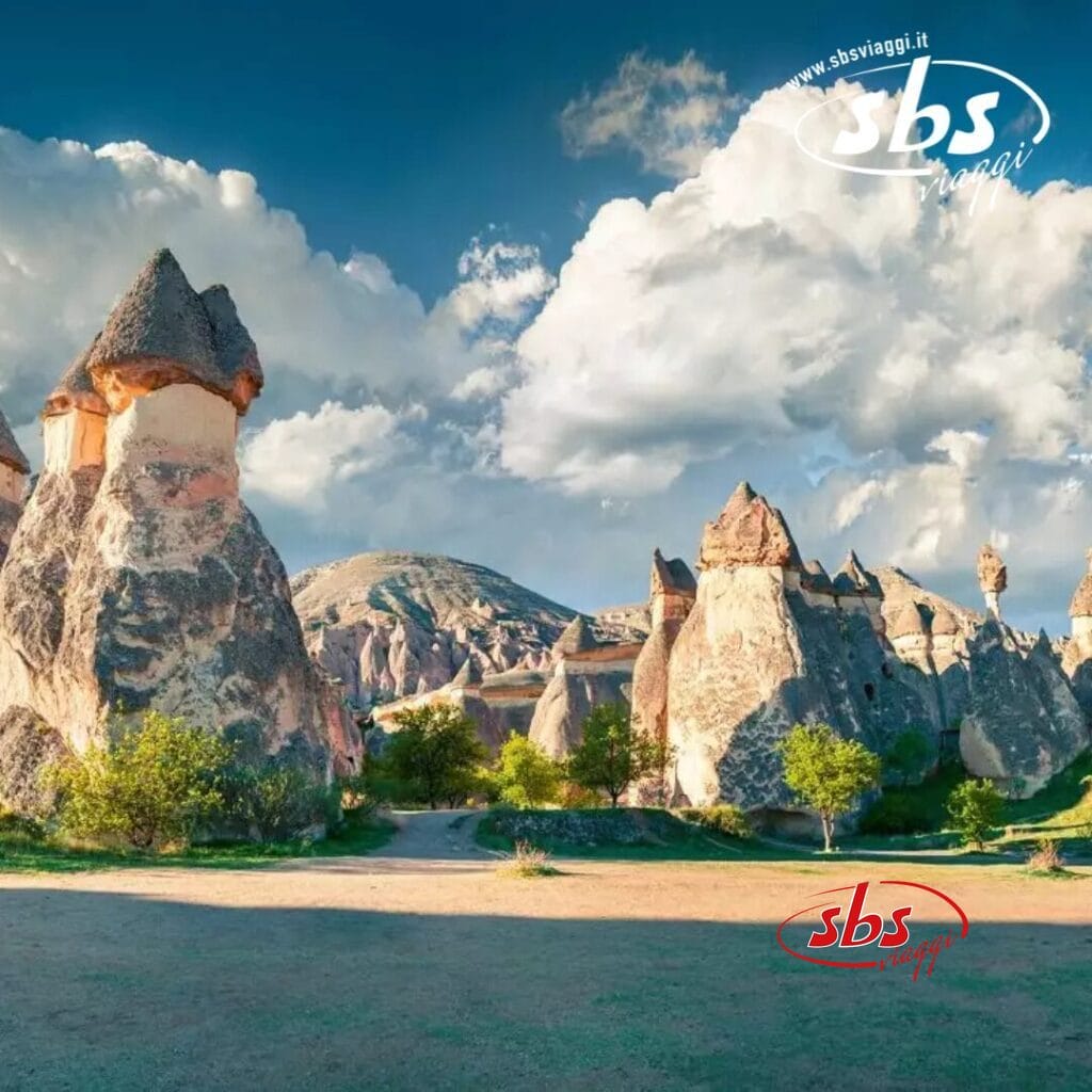 Formazioni rocciose e vegetazione sparsa sotto un cielo parzialmente nuvoloso evocano il paesaggio surreale della Cappadocia, con il logo 