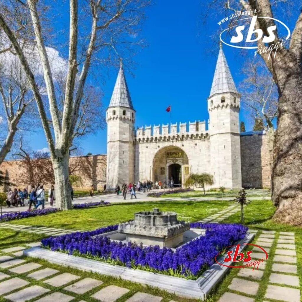 L'ingresso del Palazzo Topkapi, con le sue torri d'ingresso ornate, è incastonato tra giardini in fiore e fiori viola sotto un cielo azzurro e terso. I visitatori in un mini-tour passeggiano, immergendosi nel fascino storico di Istanbul.