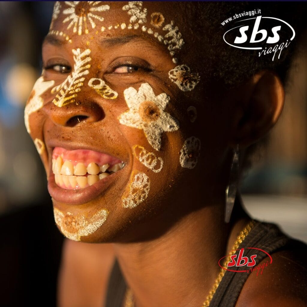 Persona sorridente con intricati disegni di pittura facciale bianca e marrone, tra cui fiori e forme geometriche, in una luce calda. La scena evoca lo spirito vibrante del Madagascar. Il logo di SBS Viaggi è nell'angolo in alto a destra.