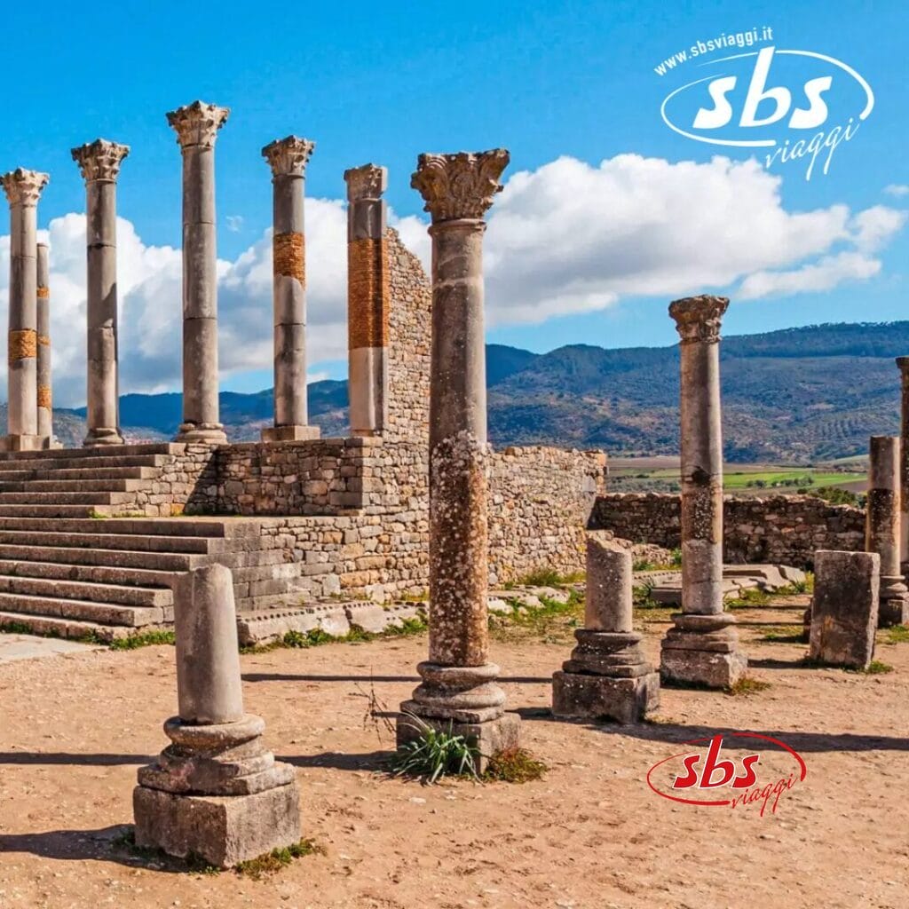 Antiche rovine con colonne e un muro parziale sotto un cielo azzurro terso, sullo sfondo di colline lontane, evocano il mistero della ricca storia del Marocco. Il logo 