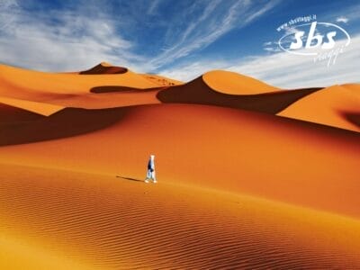 Una persona in blu passeggia nel vasto deserto del Sahara sotto un cielo parzialmente nuvoloso, con maestose dune di sabbia come sfondo. Il logo "sbs viaggi" adorna l'angolo in alto a destra, catturando il fascino di un tour di Marrakech.