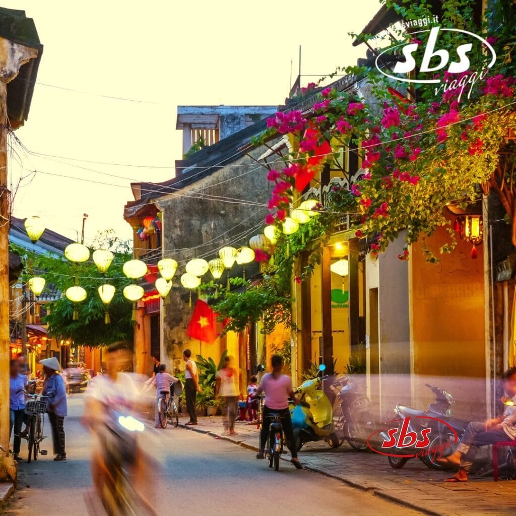 Una scena di strada vivace si svolge in Vietnam, con il movimento sfocato di persone e ciclisti. Lanterne appese in alto, mentre fiori colorati adornano gli edifici sotto un cielo serale limpido, creando un'atmosfera insolita.