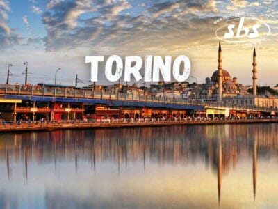 Skyline di una città con una moschea caratterizzata da due minareti in riva all'acqua, sotto un cielo nuvoloso, con sovrapposizione del testo "Tour Torino".