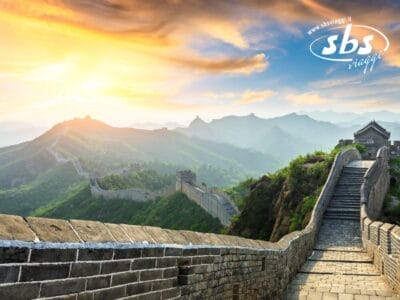 Un tratto della Grande Muraglia Cinese si snoda su verdi colline, con un tramonto vibrante e montagne sullo sfondo, evocando l'essenza incantevole dell'Antica Cina. Il logo "sbs viaggi" è elegantemente visibile nel cielo.