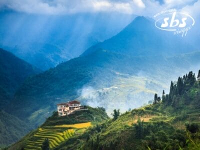 Adagiata su una collina in Vietnam, questa casa è circondata da campi terrazzati e montagne boscose sotto un cielo nuvoloso con raggi di sole che filtrano. Un logo insolito si trova elegantemente nell'angolo in alto a destra, aggiungendo un tocco di raffinatezza alla scena tranquilla.