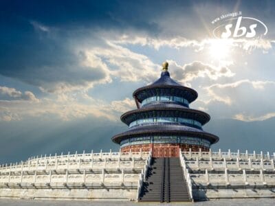 Un grande tempio decorato che ricorda quelli della Cina, con gradinate circolari e tetto blu, sorge sotto un cielo parzialmente nuvoloso, attraversato a tratti dalla luce del sole.