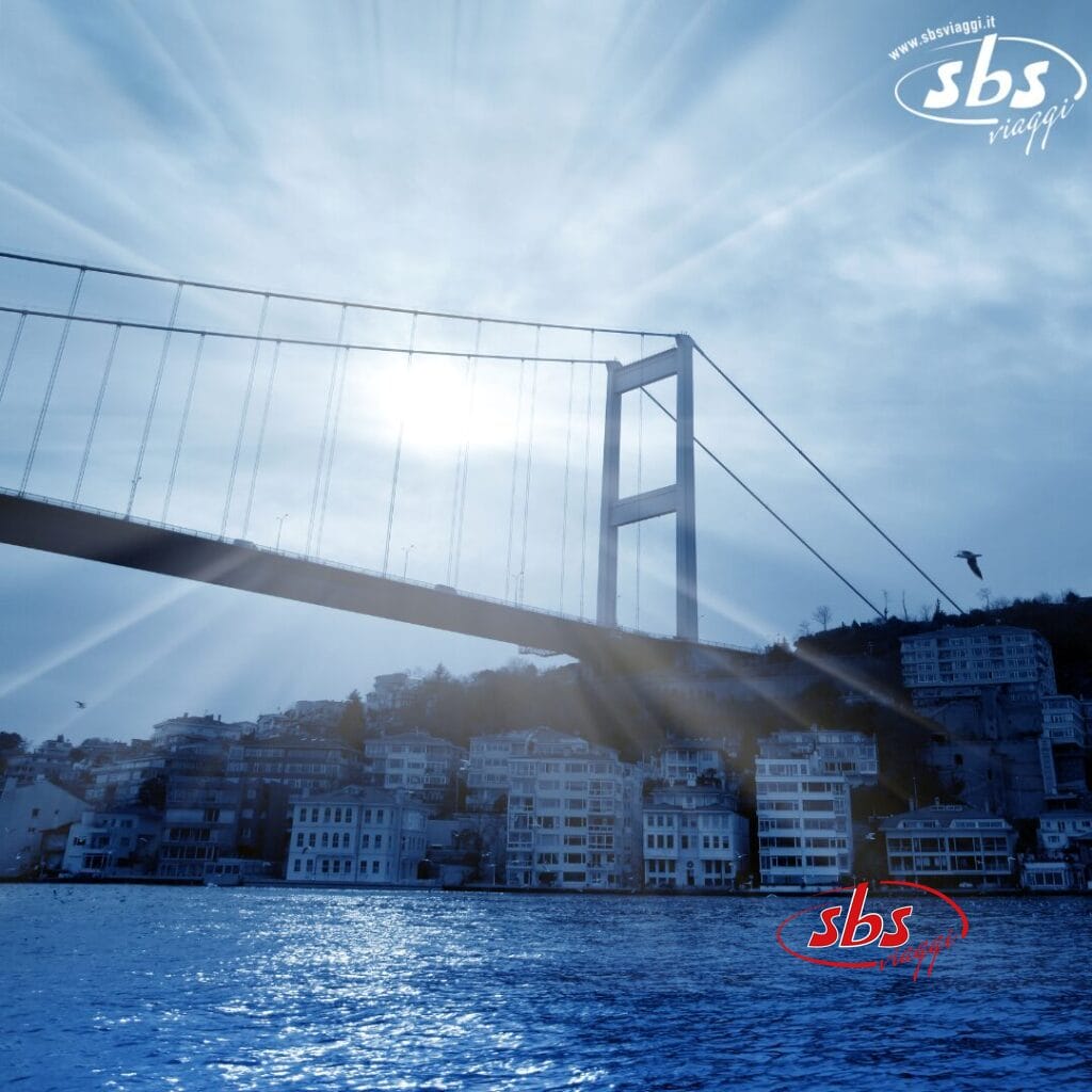 Un grande ponte sospeso si estende sopra uno specchio d'acqua, collegando il vivace paesaggio urbano di Istanbul con una collina panoramica sullo sfondo. I raggi del sole penetrano attraverso il cielo nuvoloso, offrendo uno scorcio di dove Oriente e Occidente si fondono armoniosamente.