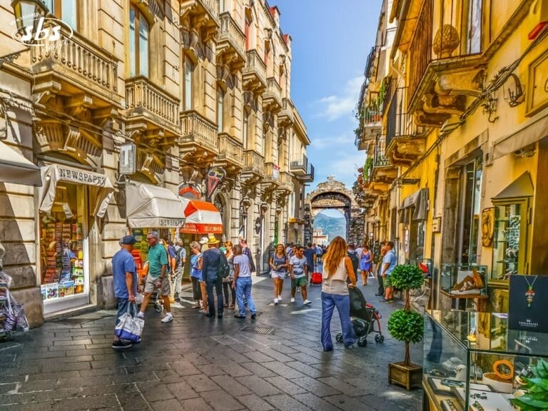 Una trafficata strada acciottolata in Sicilia, con gente che cammina, negozi su entrambi i lati, esposizioni all'aperto e un cancello ad arco in pietra visibile sullo sfondo sotto un cielo limpido.