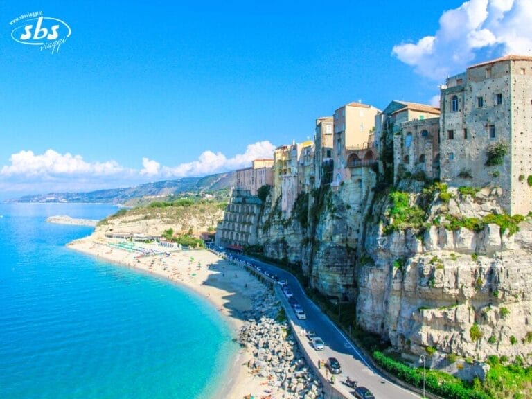 Edifici arroccati su una scogliera in Calabria si affacciano su una spiaggia sabbiosa e un mare turchese, con una strada e auto parcheggiate più in basso, sotto un cielo parzialmente nuvoloso: uno scenario mozzafiato che riecheggia l'antica bellezza della Magna Grecia.