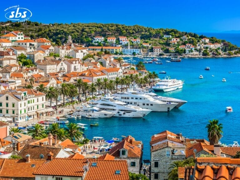 Una città costiera con edifici dai tetti rossi, palme e numerosi grandi yacht ormeggiati in un porto azzurro sotto un cielo limpido: un Incanto Mediterraneo che aspetta solo di essere scoperto durante il tuo prossimo Tour Sicilia.
