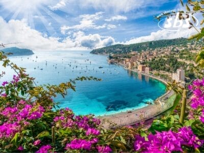 Veduta aerea di una città costiera con acque di un azzurro intenso, numerose barche e fiori viola in primo piano sotto un cielo parzialmente nuvoloso, creando una scena così pittoresca che sembra una bozza automatica uscita dal sogno di un artista.