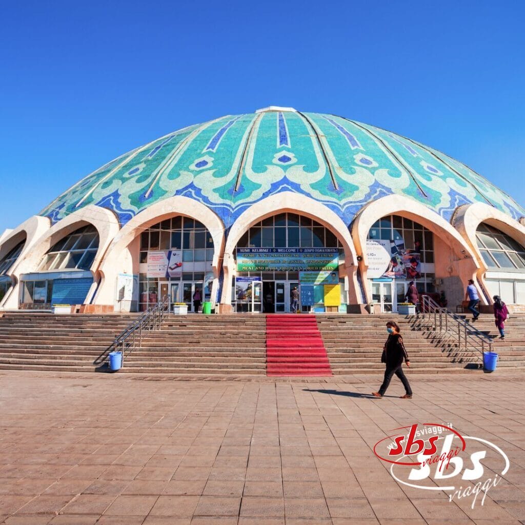 Un grande edificio a cupola con tetto decorato a mosaico, ampi gradini e ingressi ad arco si erge fiero nella Terra di Tamerlano; la gente cammina davanti sotto un cielo azzurro e terso.