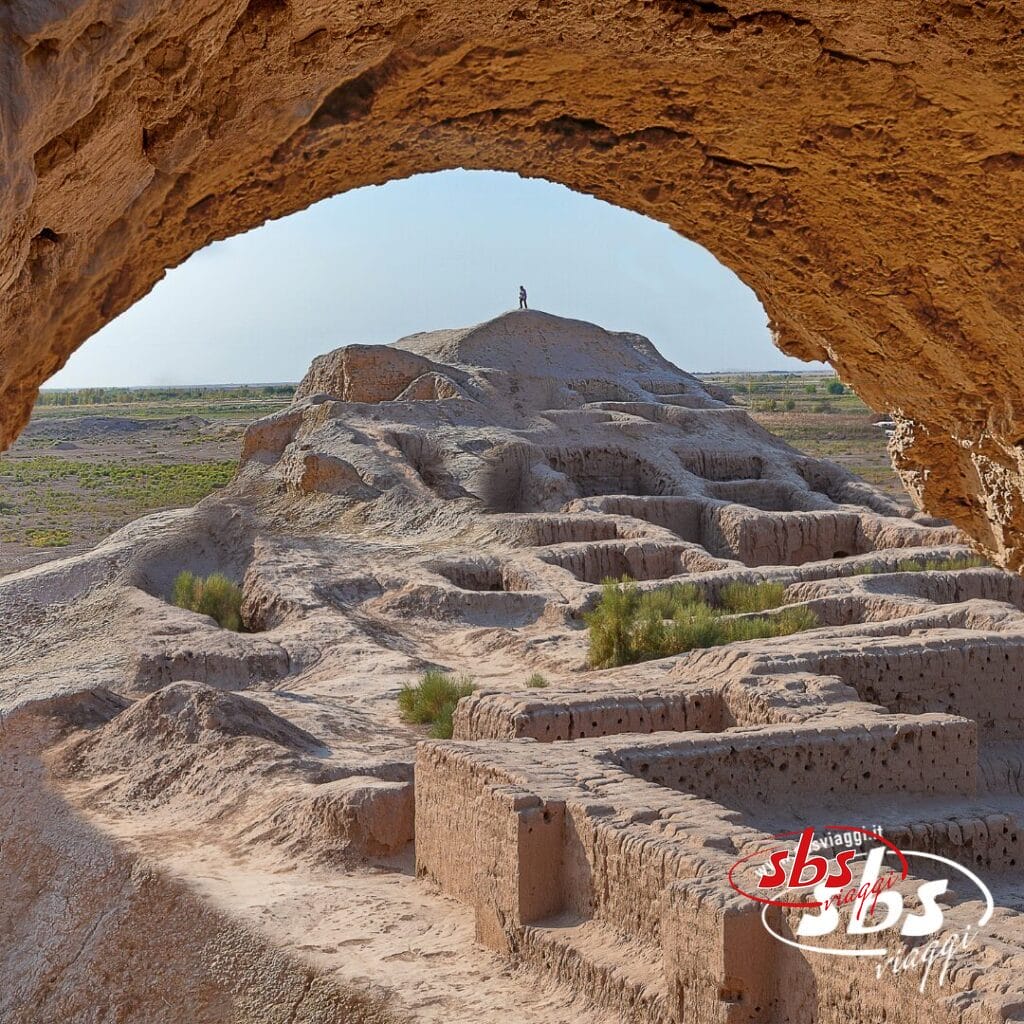 Veduta di antiche rovine e strutture stratificate in un paesaggio desertico, incorniciate da un arco roccioso, con una persona in piedi sul tumulo più alto in lontananza: echi della Terra di Tamerlano che si dispiega sotto cieli immensi.