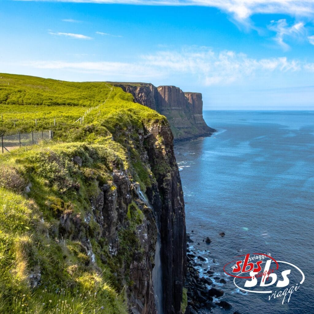 Ripide scogliere con cime erbose si affacciano su un mare calmo e azzurro sotto un cielo perlopiù limpido in Scozia. Una recinzione corre lungo il bordo della scogliera. Il logo di SBS Viaggi è visibile nell'angolo in basso a destra.