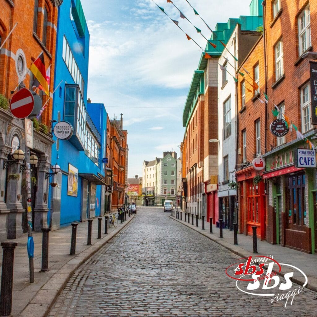 Una strada acciottolata fiancheggiata da edifici colorati, negozi e caffè, decorata con bandiere di filo sotto il limpido cielo irlandese.