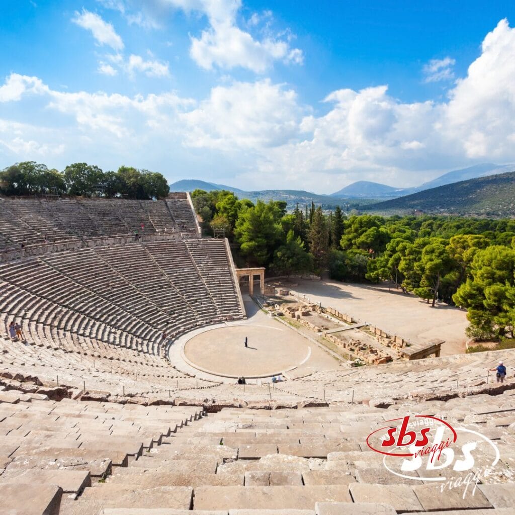 Antico anfiteatro greco con posti a sedere in pietra, circondato da alberi e montagne sotto un cielo parzialmente nuvoloso; nell'arena si vedono alcune persone, catturando la bellezza senza tempo della Grecia Classica durante il tuo prossimo tour in Grecia.