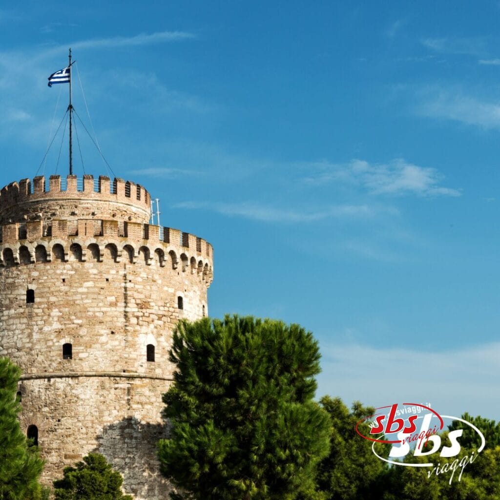 Torre rotonda in pietra con merli, con bandiera greca in cima, circondata da alberi verdi sotto un cielo azzurro e limpido a Salonicco. Scopri il fascino delle Terre di Alessandro Magno durante il tuo prossimo tour. Logo "sbs viaggi" in basso a destra.