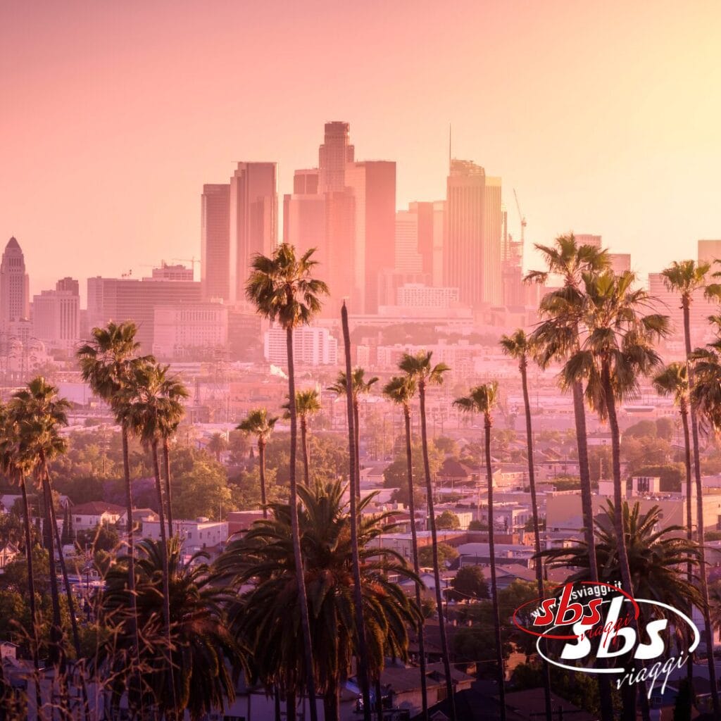 Skyline del centro di Los Angeles al tramonto con alte palme in primo piano, il logo di SBS Viaggi e un omaggio all'Explorer 1924 nell'angolo in basso a destra.