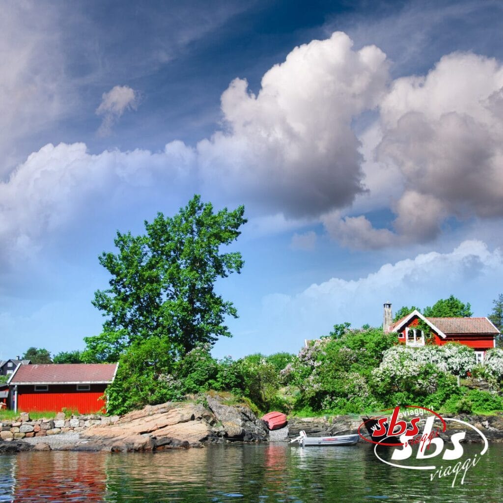 Una piccola casa rossa con dependance si trova su una lussureggiante costa rocciosa nella Terra dei Fiordi, in Norvegia, con barche nelle vicinanze sotto un cielo azzurro costellato di grandi nuvole. Il logo "sbs viaggi" è visibile in basso a destra.
