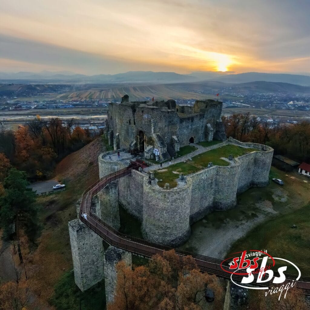 Vista aerea delle rovine di un grande castello in pietra circondato da alberi, con un tramonto sui campi e sulle colline sullo sfondo: uno spettacolo indimenticabile per chi visita la Romania.