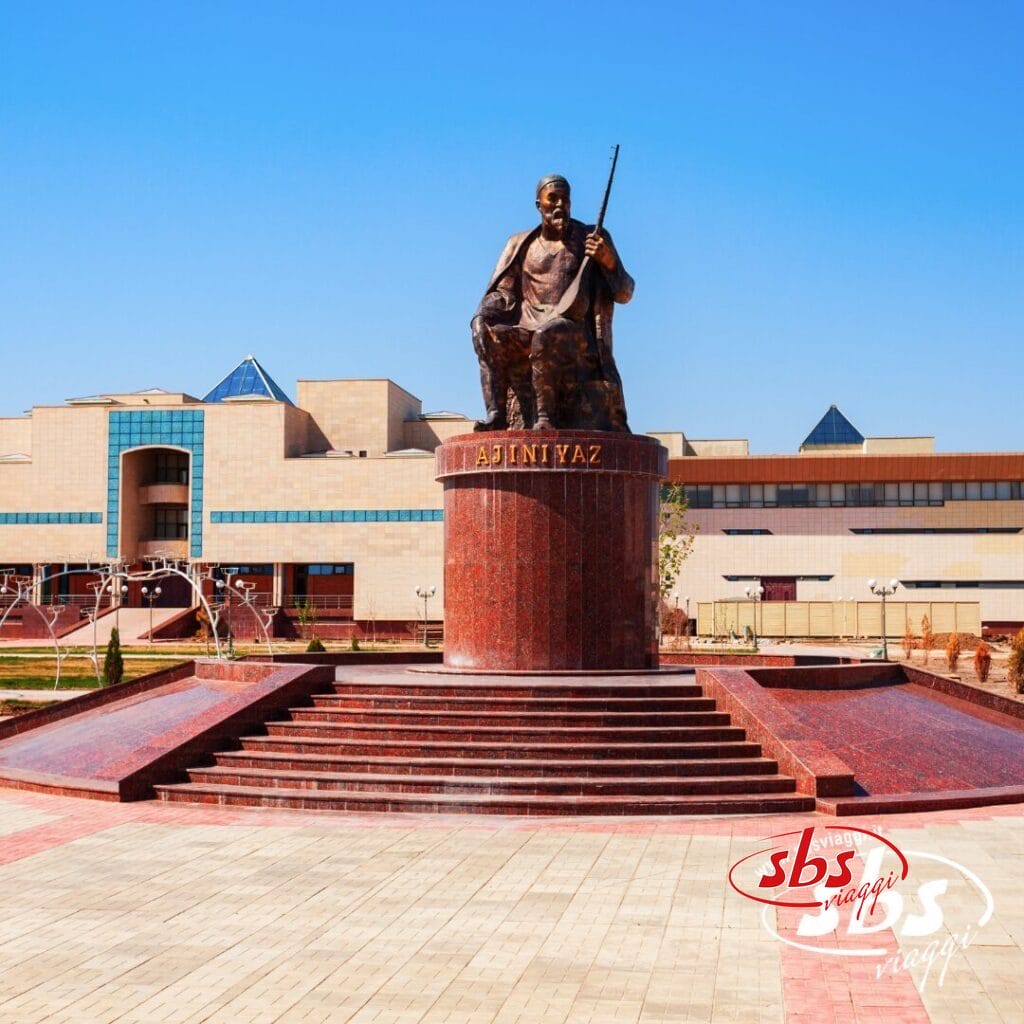 Una grande statua in bronzo raffigurante un uomo seduto con in mano un fucile, montata su un piedistallo di marmo rosso, si erge di fronte a un edificio moderno sotto il limpido cielo azzurro della Terra di Tamerlano, vicino al Lago d'Aral.