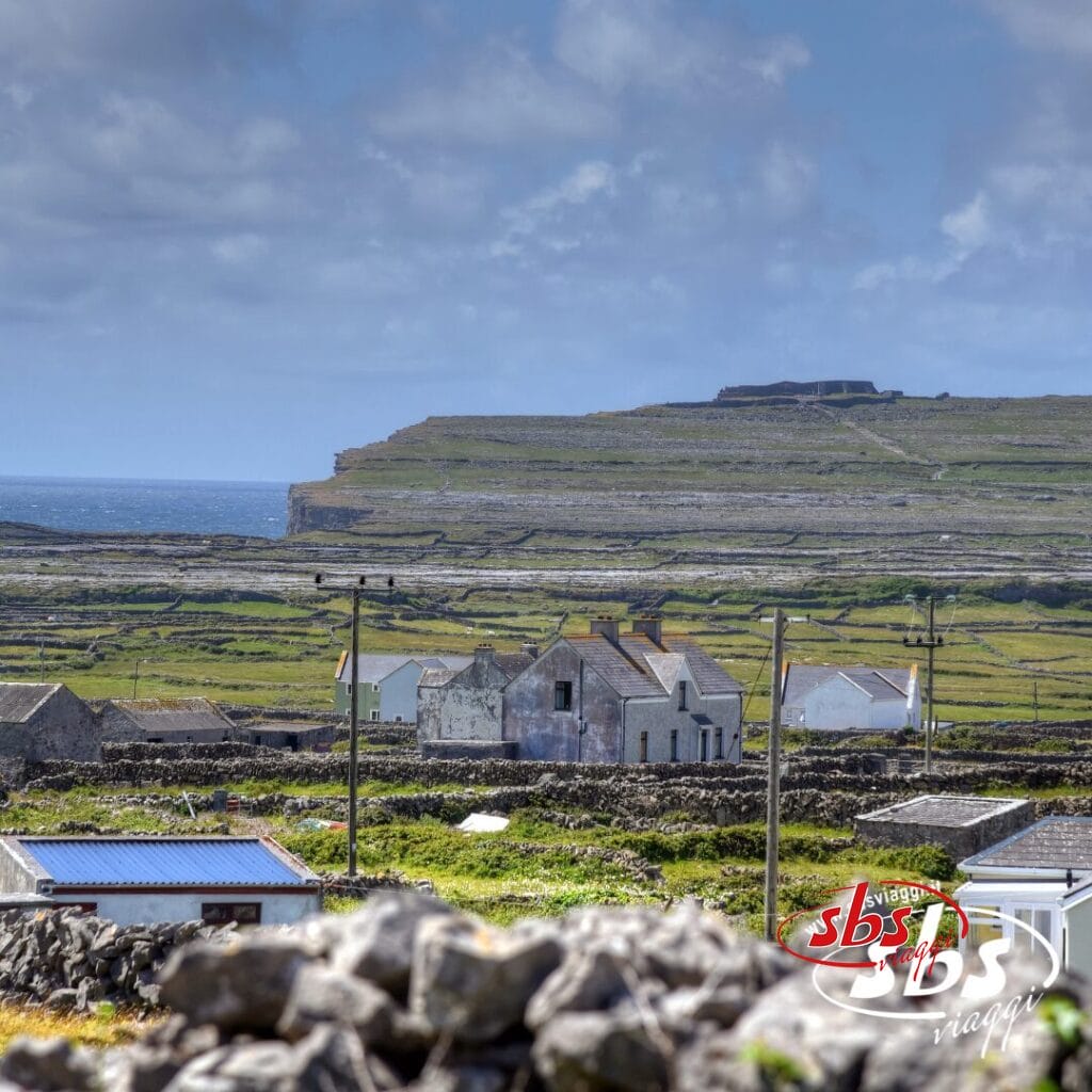 Un paesaggio rurale nelle Isole Aran in Irlanda, caratterizzato da case in pietra, muri in pietra e campi verdi sotto un cielo nuvoloso, vicino a una costa frastagliata.
