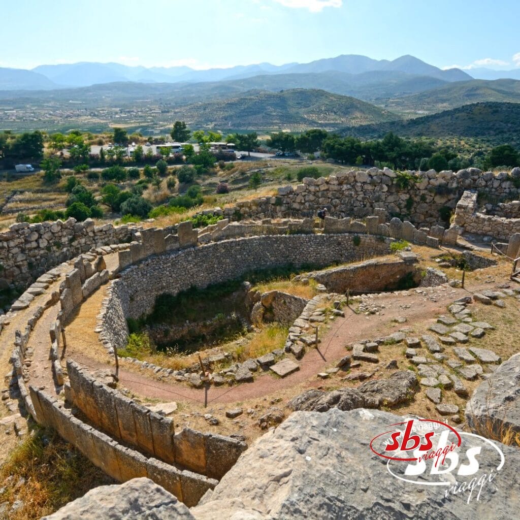 Antiche rovine in pietra di una struttura circolare si trovano su un pendio, circondate da montagne e vegetazione sotto un cielo limpido: uno scenario iconico per qualsiasi avventura Viaggio Grecia o Tour Grecia.