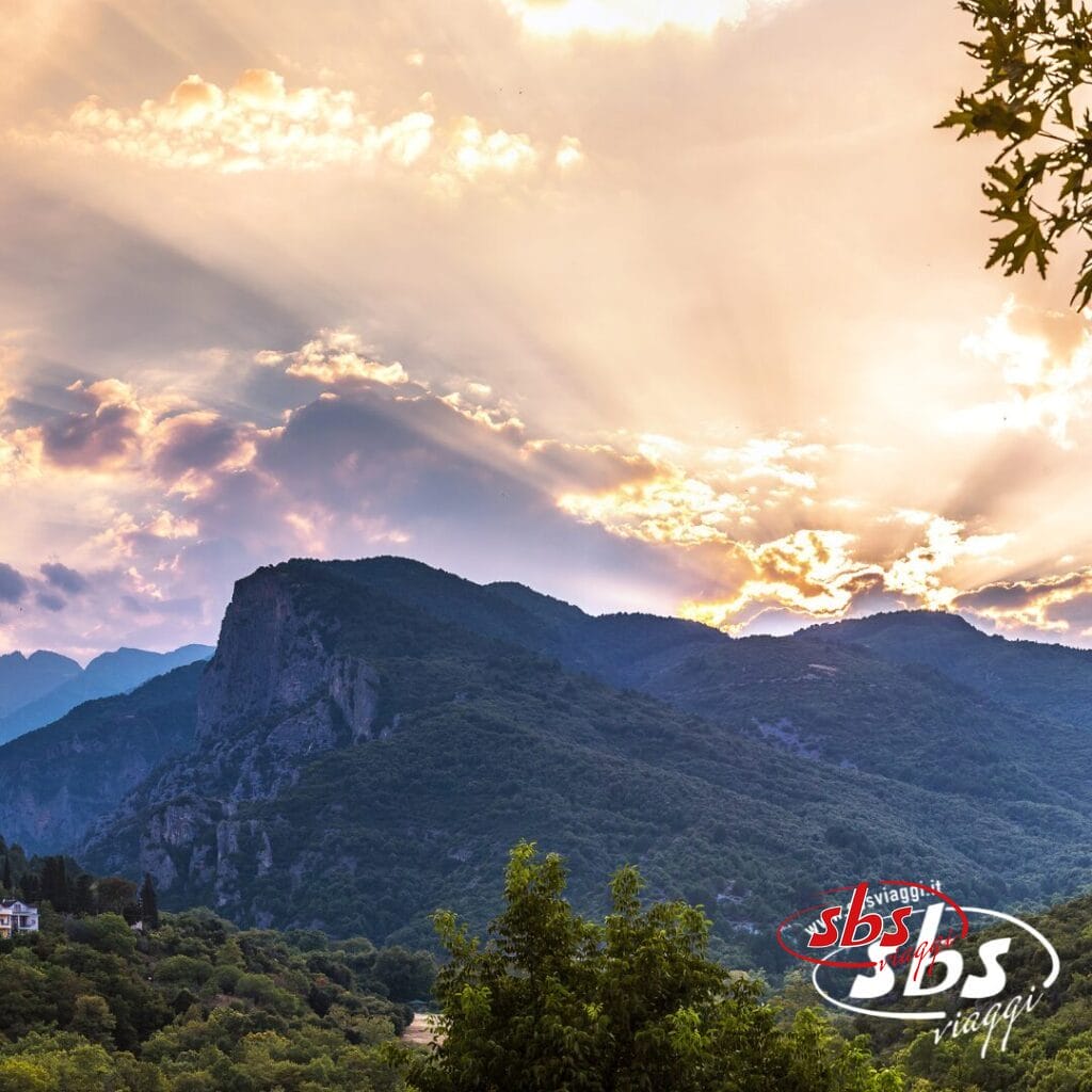 La luce del sole filtra tra le nuvole su un paesaggio montuoso dalla vegetazione lussureggiante, evocando la bellezza delle Terre di Alessandro Magno. Il logo 