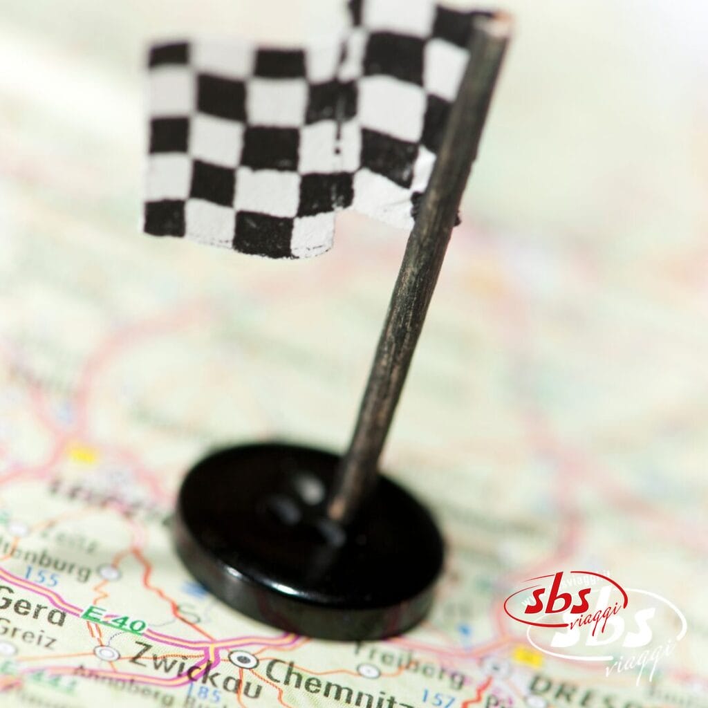 Una piccola spilla a forma di bandiera a scacchi indica un punto sulla mappa, con città e strade visibili sullo sfondo: perfetta per seguire il Grand Tour del 2025 o pianificare le tue avventure in MotoGP.