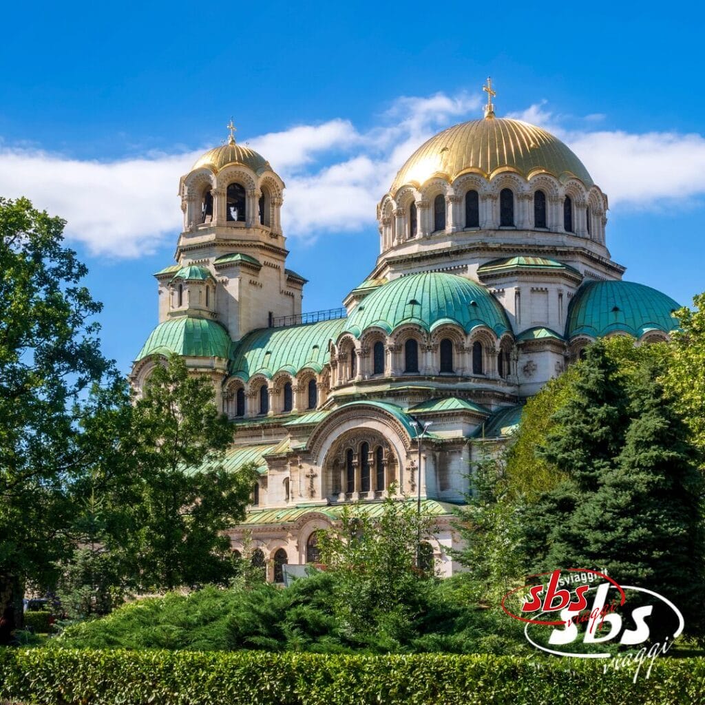 Una grande cattedrale con cupole verdi e tetti dorati si erge dietro gli alberi sotto un cielo azzurro: uno spettacolo mozzafiato durante un'avventura del Tour Bulgaria; il logo 