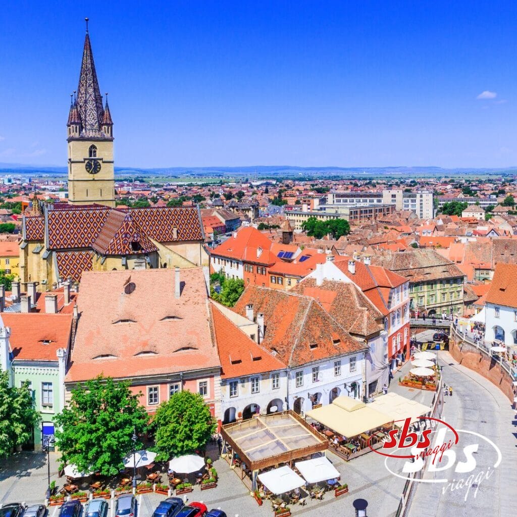 Vista aerea di una piazza cittadina europea in Romania, con caffè all'aperto, edifici dai tetti rossi, una chiesa con tetto decorato e una torre dell'orologio sotto un cielo limpido: perfetta per chi desidera visitare la Romania.