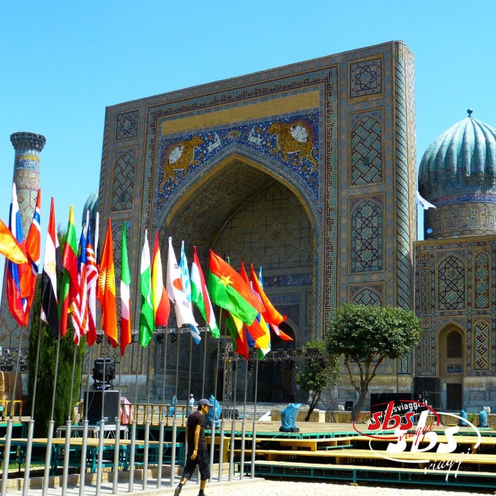 Uno storico edificio architettonico islamico con intricate decorazioni in piastrelle, un grande ingresso ad arco e numerose bandiere nazionali esposte sulla facciata, che ricordano la magnificenza della Terra di Tamerlano.