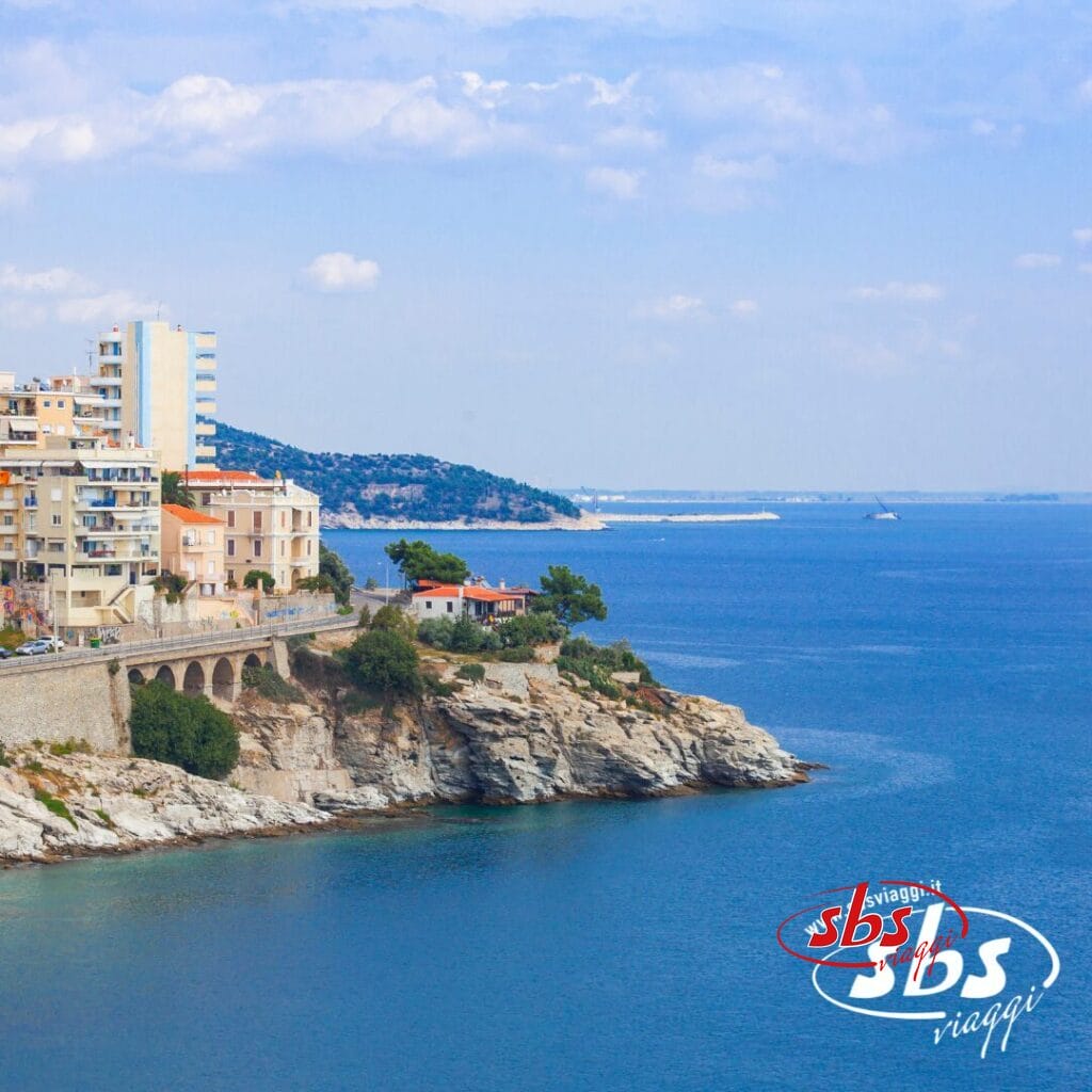 Paesaggio urbano costiero con edifici a picco sul mare azzurro sotto un cielo parzialmente nuvoloso; scopri Salonicco, il gioiello delle Terre di Alessandro Magno, nel tuo prossimo tour. Logo SBS Viaggi nell'angolo.