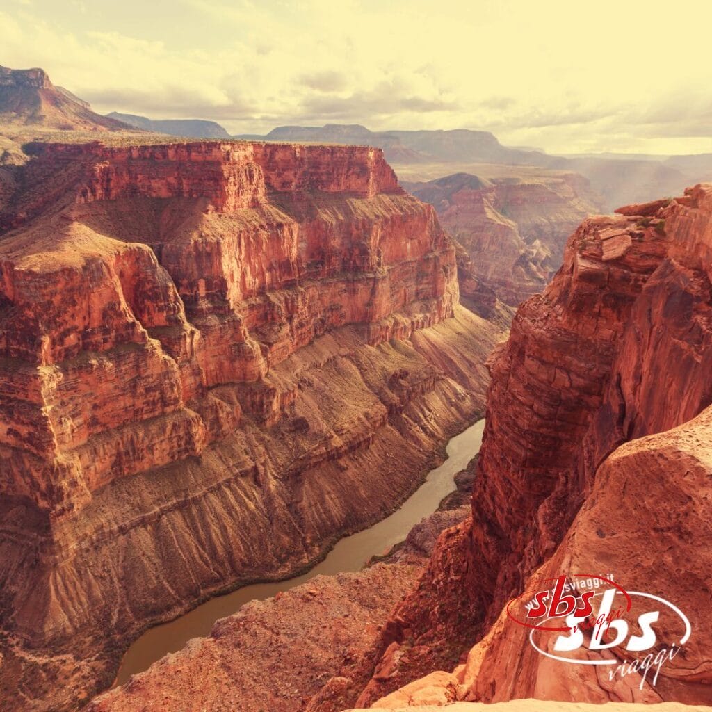 Un'ampia visuale del Grand Canyon mostra scogliere di roccia rossa stratificate e un fiume che serpeggia attraverso il fondo del canyon sotto un cielo limpido, che ricorda i paesaggi di Los Angeles immortalati nel 1924.