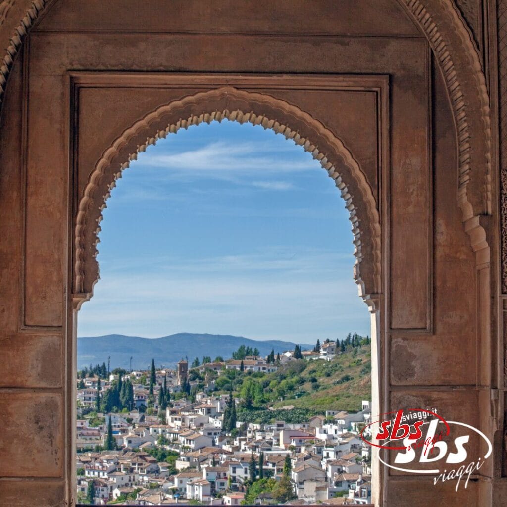 Vista attraverso un arco decorato che mostra una pittoresca cittadina andalusa con edifici bianchi, alberi verdi e dolci colline sotto un cielo azzurro: un pittoresco invito a visitare questa incantevole regione.