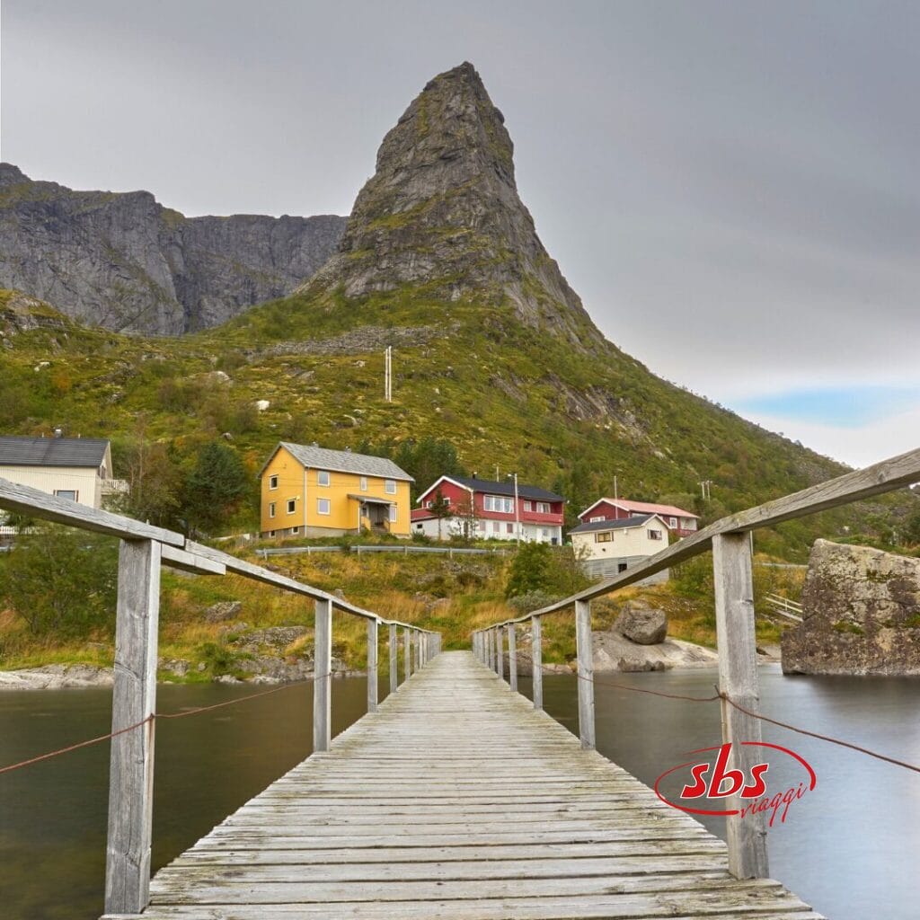Una passerella di legno conduce a un piccolo gruppo di case colorate ai piedi di una ripida montagna rocciosa, sotto un cielo coperto, nell'incantevole Terra dei Fiordi, in Norvegia.