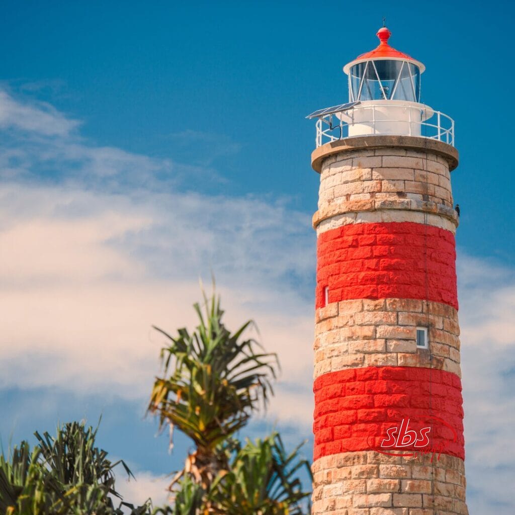 Un faro in pietra con strisce orizzontali rosse e bianche si erge accanto a piante verdi e rigogliose sotto un cielo azzurro con nuvole sparse, evocando la bellezza paesaggistica di un tour delle Lofoten.