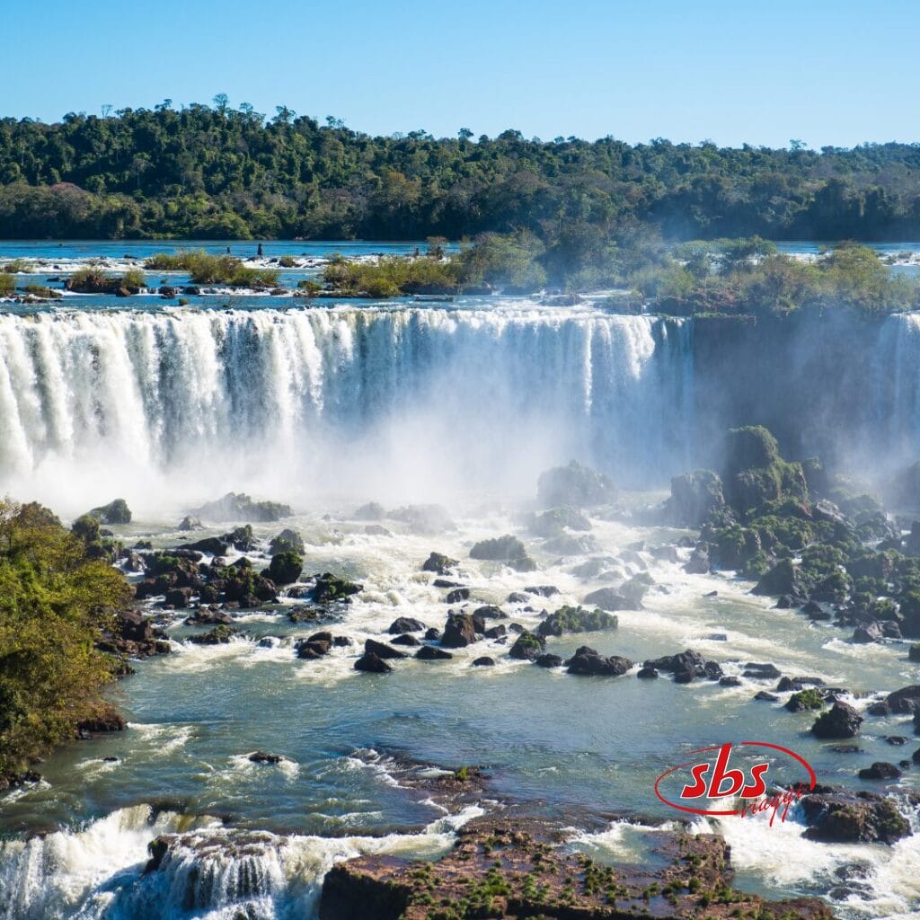 Un'ampia cascata si riversa da una parete rocciosa nel fiume sottostante, mentre la nebbia si alza e sullo sfondo si estende una fitta foresta sudamericana, sotto un cielo limpido.