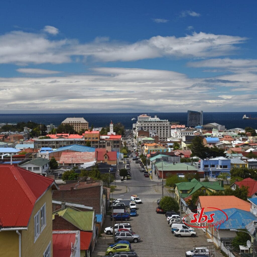 Un paesaggio urbano costiero che ricorda la Patagonia, con edifici colorati, auto parcheggiate e una strada dritta che conduce al mare sotto un cielo azzurro con nuvole sparse.