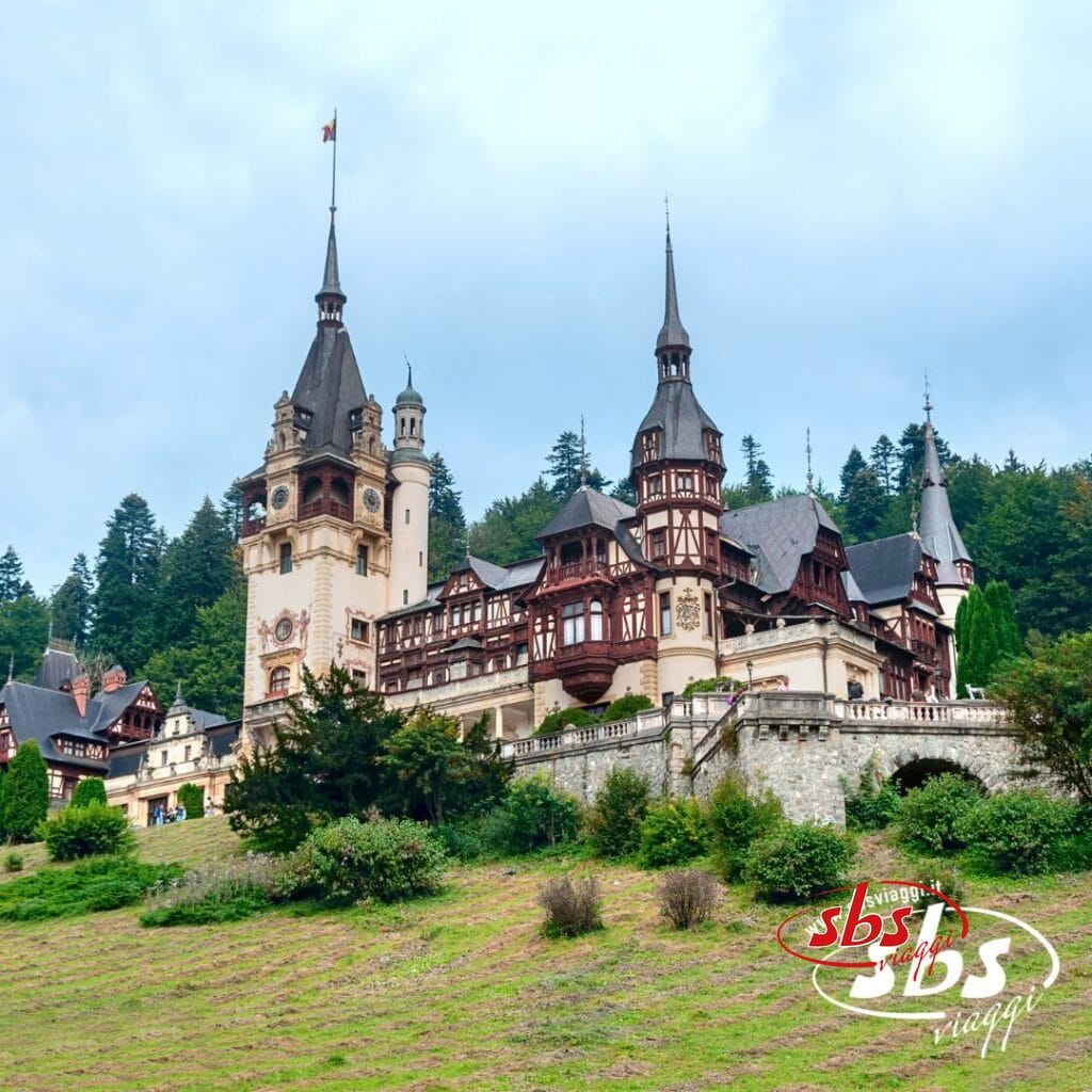 Il Castello di Peles, con le sue torri ornate, sorge su un pendio erboso in Romania, circondato da alberi sotto un cielo nuvoloso. Il logo 