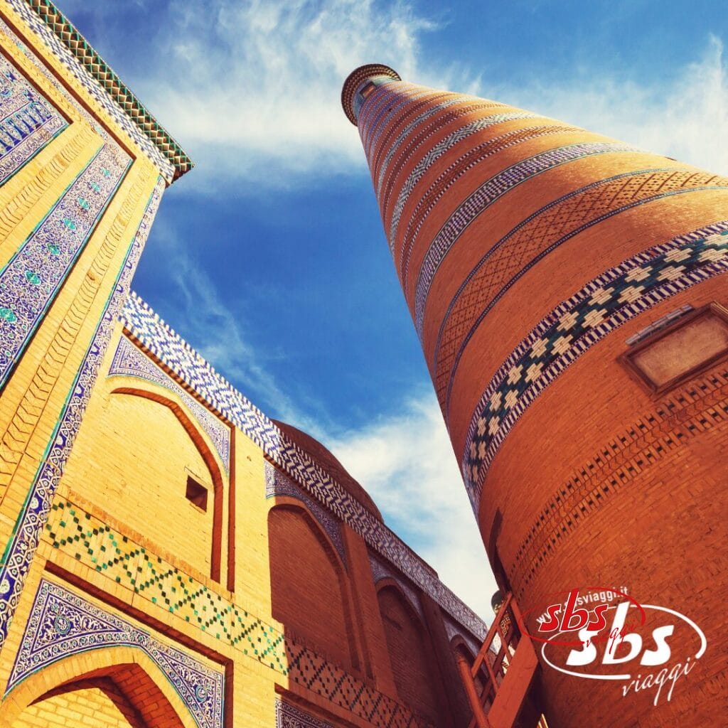 Un alto minareto cilindrico in mattoni con motivi geometrici blu si erge accanto a un edificio riccamente piastrellato sotto un cielo parzialmente nuvoloso nella leggendaria Terra di Tamerlano. Il logo