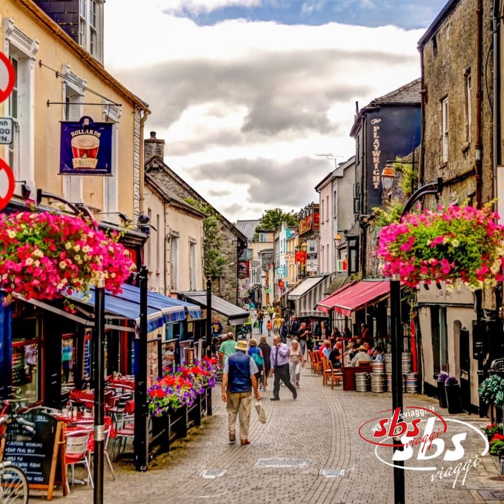 Una strada acciottolata fiancheggiata da negozi, ristoranti e cesti di fiori appesi; la gente passeggia nell'area pedonale sotto Cieli d'Irlanda, l'incantevole cielo irlandese.