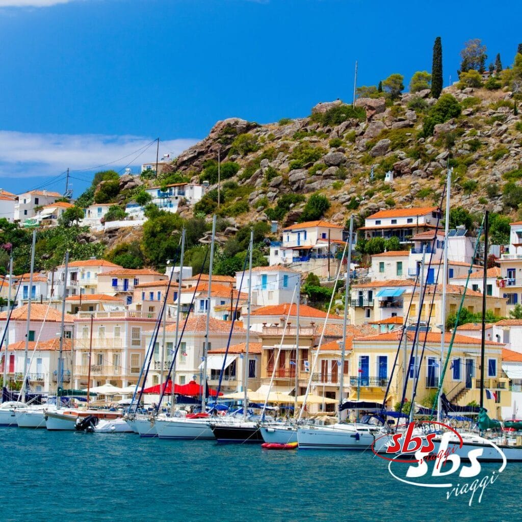 Case colorate con tetti arancioni fiancheggiano una collina dietro un porto turistico pieno di barche a vela, catturando il fascino di un'avventura del Tour Grecia. Il logo di SBS Viaggi è visibile nell'angolo in basso a destra.